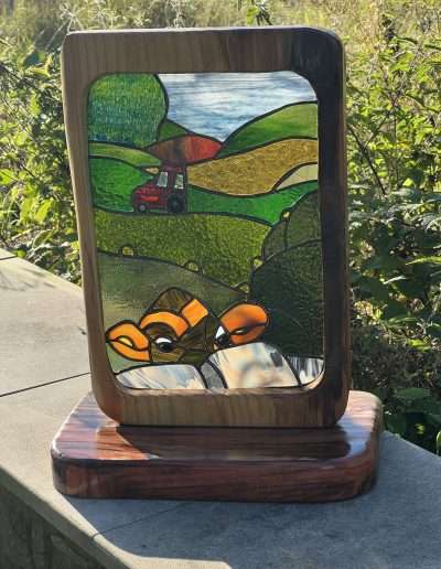 Stained glass and wood ornament of a cow looking over a stone wall with a tractor in the background